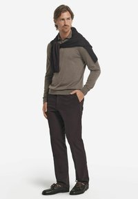 Brun långärmad tröja med rund halsringning, kombinerad med mörka byxor och svarta skor, med en svart tröja draperad över axlarna.