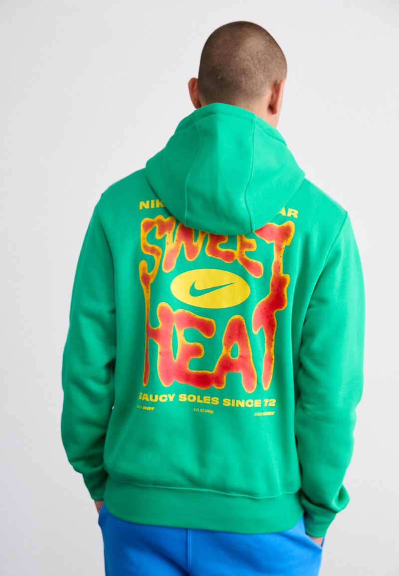 Grøn hættetrøje med et stort, flerfarvet "SWEET HEAT" grafik på ryggen, præget af en gul Nike swoosh. Ribstrikkede ærmer og kant.