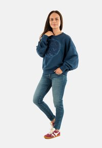 Sudadera azul marino de gran tamaño con un diseño gráfico circular, combinada con jeans ajustados de color azul claro y zapatillas color burdeos con detalles en blanco.