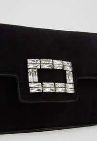 Clutch de ante negro con detalle de hebilla rectangular de cristal plateado en la solapa frontal sobre un fondo liso.