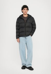 Schwarze Daunenjacke mit Reißverschluss, verfügt über eine Kapuze und gepolsterte Einsätze. Getragen über einem braunen Shirt und kombiniert mit hellblauen, weiten Jeans.