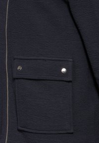 Giacca strutturata blu navy, con chiusura a zip e tasca frontale con dettagli in metallo argentato. Il tessuto presenta un leggero motivo a righe.