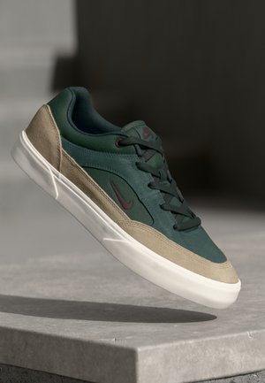 Vihreä ja beige Nike-tennari valkoisella pohjalla ja tummilla nauhoilla, roikkumassa betonipinnan yläpuolella tumman taustan edessä.