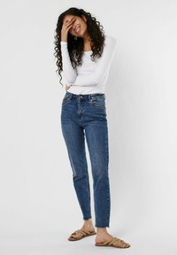 Hochgeschnittene blaue Jeans aus Denim mit leichtem Stretch, ausgestattet mit einem Fünf-Taschen-Design und rohen Säumen, kombiniert mit einem weißen Langarmoberteil.