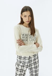 Beige sweatshirt med sort tekst, der siger "MERE SØVN MERE KRAM MERE KÆRLIGHED," parret med ternede pyjamasbukser i sort og hvid.