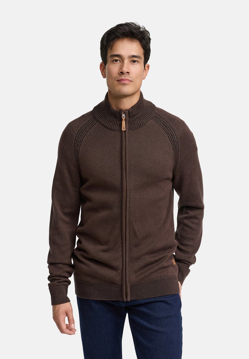 Brązowy dziany sweter z wysokim kołnierzem, zapinany na zamek, z prążkowanymi detalami, zestawiony z ciemnoniebieskimi dżinsami. Miękka tekstura i dopasowany fason.