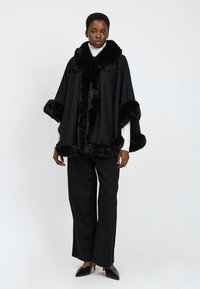Donna che indossa un mantello nero con bordi in pelliccia, un dolcevita bianco, pantaloni larghi neri e décolleté a punta nera su uno sfondo bianco.