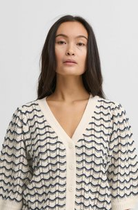 Cardigan crème avec un motif en vagues bleu marine, décolleté en V, manches courtes et trois boutons sur le devant. Design tricoté texturé.