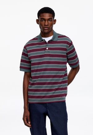 Man draagt een korte mouw bordeauxrode en grijze gestreepte poloshirt met marineblauwe broek, staand tegen een effen witte achtergrond.