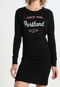 Svart långärmad klänning med grafiken "Forest Park Portland 1994" i vitt och rosa. Mjuk bomullsmaterial, figursydd design, ovanför knäna.
