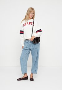 Biała bluza z czerwonym napisem "GAP NYC", z granatowymi i czerwonymi paskami na rękawach, w zestawie z jasnoniebieskimi luźnymi dżinsami i błyszczącymi ciemnoczerwonymi balerinami.