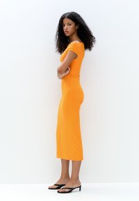 Robe midi ajustée orange avec des manches courtes et un design de couture latérale, associée à des sandales flip-flop noires, sur un fond blanc.