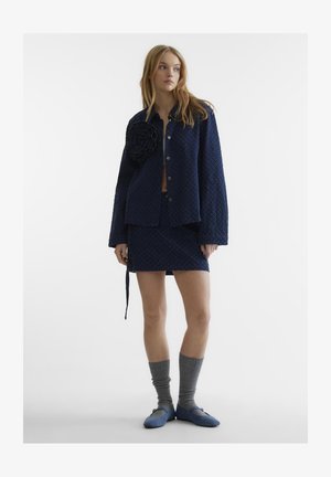 Jachetă texturată de culoare bleumarin cu un accent floral mare, asociată cu o fustă asortată, ciorapi gri până la genunchi și pantofi plat albastri.