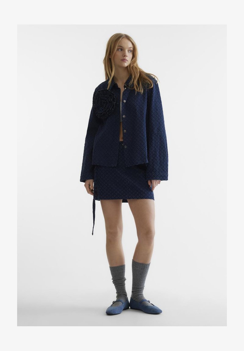 Μπλε υφασμάτινο σακάκι με μεγάλο floral σχέδιο, συνδυασμένο με μίνι φούστα, γκρι γόνατες κάλτσες και μπλε φλατ παπούτσια.