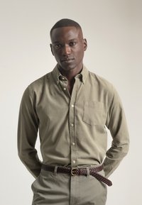 Camisa de botonada verde oliva con un bolsillo en el pecho, de material de algodón, combinada con pantalones gris claro y un cinturón trenzado marrón. Textura suave.