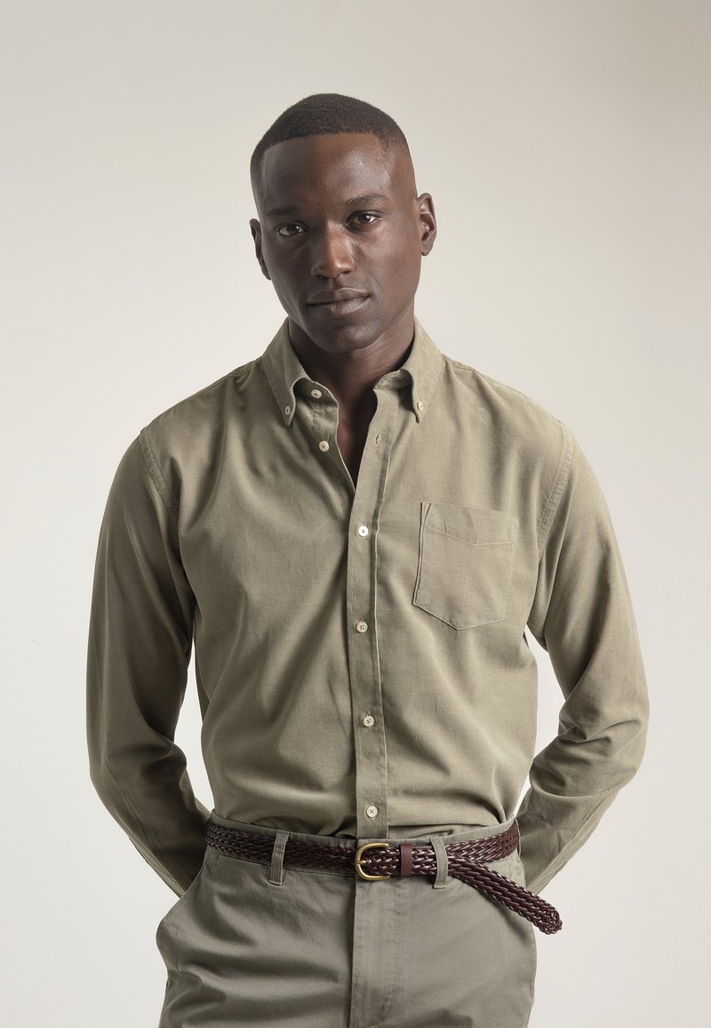 Camisa de botonada verde oliva con un bolsillo en el pecho, de material de algodón, combinada con pantalones gris claro y un cinturón trenzado marrón. Textura suave.