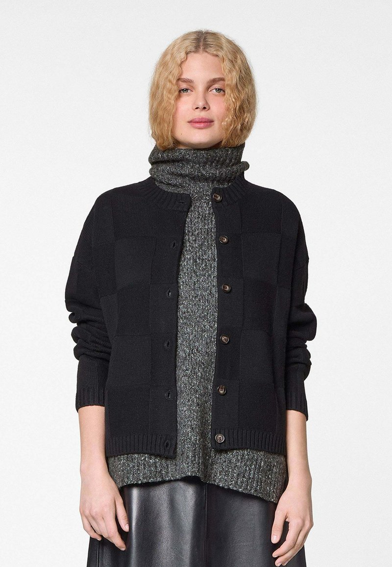 Zwarte cardigan met een gestructureerd checkpatroon, voorzien van een geribbelde kraag en manchetten, met knoopsluiting, gelaagd over een grijze coltrui.