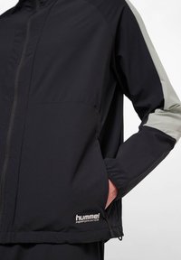Svart lättviktsjacka med grå detaljer på ärmarna, har en framdragkedja, sidofickor och "hummel PERFORMANCE"-logotyp på fållen.
