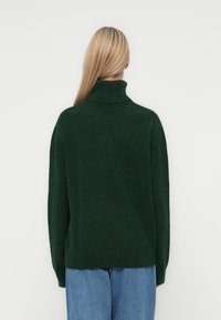 Pull col roulé en tricot texturé vert foncé avec une coupe décontractée et un ourlet côtelé, vu de dos. Associé à un jean en denim bleu clair.