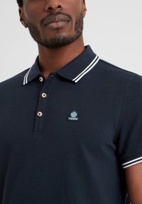 Navy polo shirt with white striped collar, three-button placket, and a small blue and white logo on the chest. Textured fabric.