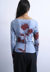 Femme aux longs cheveux foncés portant un haut à manches longues bleu clair avec de grandes imprimés floraux rouge foncé et un pantalon noir, vue de dos.