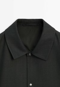 Veste noire à col avec fermeture par boutons et motif discret à fines rayures verticales sur le tissu.
