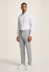 Mand, der står iført en hvid skjorte med knapper, lysegrå smalle bukser og hvide sneakers foran en ensfarvet baggrund.