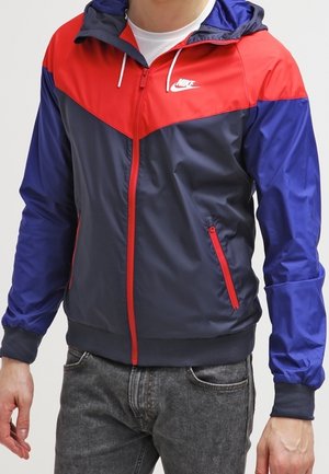 Mężczyzna w czerwono-granatowo-niebieskiej kurtce przeciwdeszczowej z kapturem marki Nike, z czerwonym zamkiem błyskawicznym i kieszeniami, w połączeniu z szarymi dżinsami.