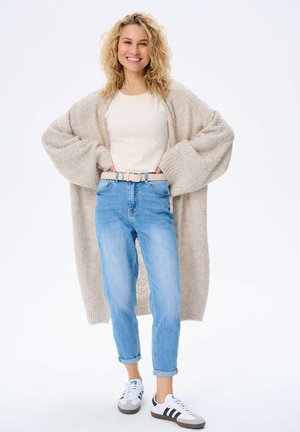 Beige knitted cardigan, cream top, light blue jeans, and white sneakers with black stripes. Model has curly hair and is smiling.