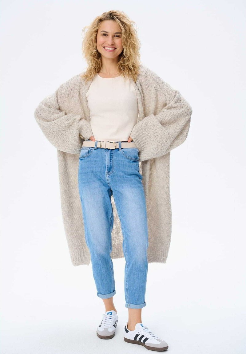 Beige gebreide cardigan, crèmekleurig top, lichtblauwe jeans en witte sneakers met zwarte strepen. Model heeft krullend haar en lacht.