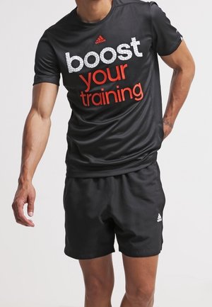 Mężczyzna w czarnym sportowym stroju Adidas z napisem „boost your training” na koszulce, stojący na jasnym jednolitym tle.
