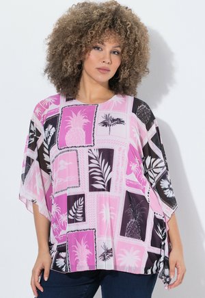 Mujer con cabello rizado que lleva una camiseta rosa holgada con patrones tropicales y de piña en negro y blanco, de pie sobre un fondo blanco liso.