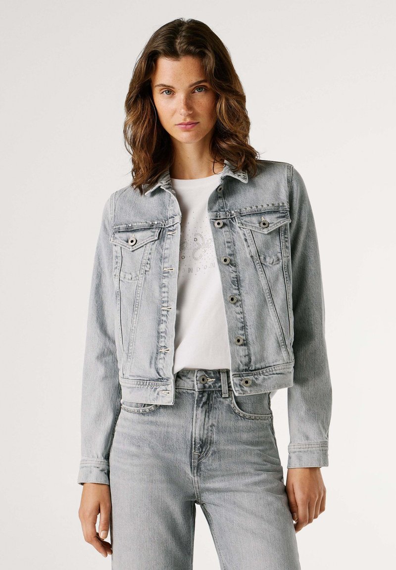 Licht gewaschene Denimjacke mit Knopfverschluss, zwei Brusttaschen und einem cropped Design. Getragen über einem weißen Grafik-T-Shirt und passenden Jeans.