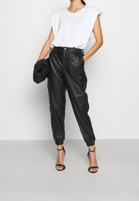 Person som bär en vit ärmlös topp, svarta högmidjade läderjoggingbyxor, svarta remsandaler med klack och håller en svart läderväska.