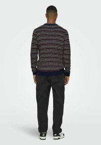 Pull en tricot avec un motif géométrique bleu marine et rouge, col rond, poignets et ourlet côtelés ; associé à un pantalon cargo noir et des baskets.
