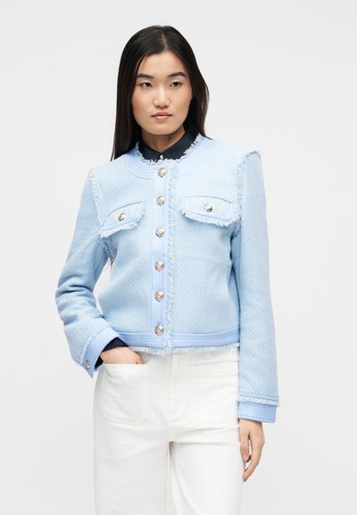 Γυναίκα που φοράει  γαλάζιο υφασμάτινο σακάκι με κουμπιά και σκισμένα τελειώματα, και λευκό παντελόνι, στέκεται με το ένα χέρι στην τσέπη μπροστά σε λευκό φόντο.