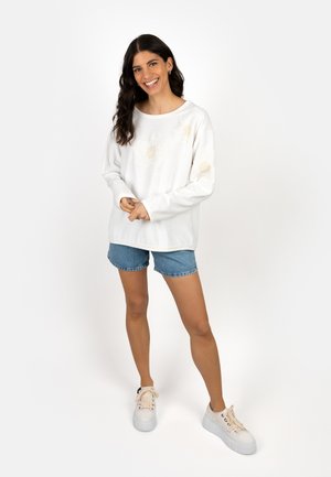 Smiling woman stands with hands clasped, wearing white long-sleeve shirt, denim shorts, and white platform sneakers on white background.