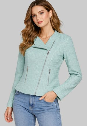 Femme portant une veste zippée texturée bleu clair avec des détails de fermeture éclair diagonale, associée à un jean bleu clair, debout devant un fond uni.