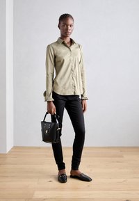 Camisa verde oliva de botões com mangas volumosas, calças pretas de corte justo e uma bolsa preta. Mocasins pretos com detalhes em metal completam o visual.