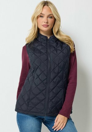 Femme portant un gilet matelassé noir par-dessus un haut à manches longues bordeaux et un jean bleu, debout devant un fond clair uni.