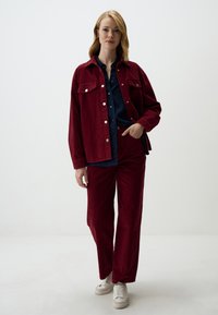 Rode corduroy jas met zilveren knopen en zakken, gecombineerd met bijpassende broeken. De outfit wordt aangevuld met een marineblauw shirt eronder en witte sneakers.