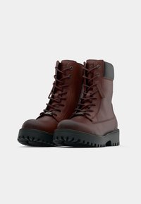 Par de botas de couro castanho com atacadores, solas grossas pretas e golas almofadadas no tornozelo, em fundo branco.