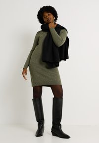 Vestido de punto verde con mangas largas y dobladillo acanalado. Combinado con una bufanda negra, la modelo lleva botas altas negras, acentuando el conjunto.