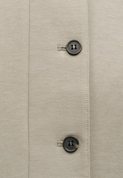 Bež pletena tkanina z navpičnim šivom in dvema temnosivima, okroglih gumbov s štirimi luknjicami, všitima v eno samo kolono.