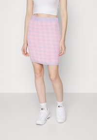 Mujer con una falda de pata de gallo a media pierna en tonos rosa y lavanda, top blanco corto, calcetines blancos y zapatillas blancas, de pie contra un fondo blanco.