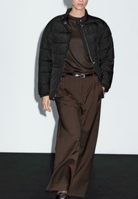 Person walking, wearing black puffer jacket over dark brown top and wide-legged brown trousers with black belt, hands partially in pockets.