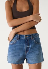 Keskisinis denim-lyhythousut, joissa on revitty hame, korkea vyötärö ja viisi taskua. Ominaisuudet sisältävät kontrastiväriset saumat ja etupainon kiinnityksen.