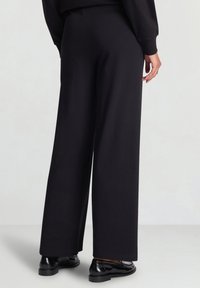 Hose mit weitem Bein aus schwarzem Stoff mit glatter Textur; hoher Bund und wenige Nähte; kombiniert mit polierten schwarzen Schuhen.