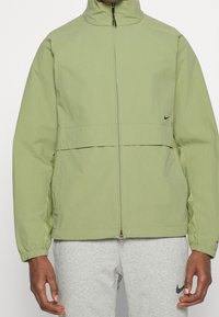 Chaqueta verde claro con cremallera, cuello alto, puños elásticos y una costura horizontal en la cintura. Presenta un pequeño logotipo negro en el lado izquierdo del pecho.
