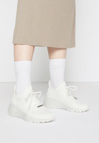 White athletic sneakers with a textured mesh upper, lace-up design, and raised rubber sole, paired with white crew socks and a beige ribbed skirt.